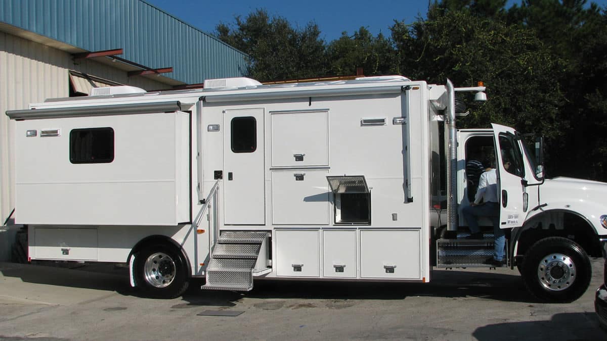 Large white mobile command center vehicle parked outdoors, featuring numerous compartments, windows, and a side door with steps.