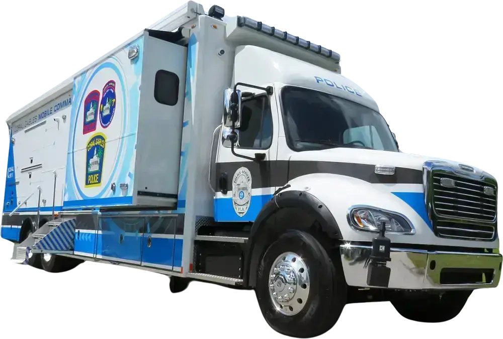 A large white and blue Coral Gables Police Mobile Command vehicle is parked on a flat surface. The vehicle features official police logos, side compartments, a front cab, and emergency lights mounted on the roof.
