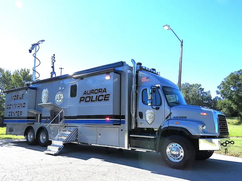 A large Aurora Police mobile command center truck is parked outdoors on a sunny day. The vehicle has multiple wheels, steps leading to a door, and equipment including a raised antenna on its roof. Trees and grass are in the background. Aurora Police Mobile Command