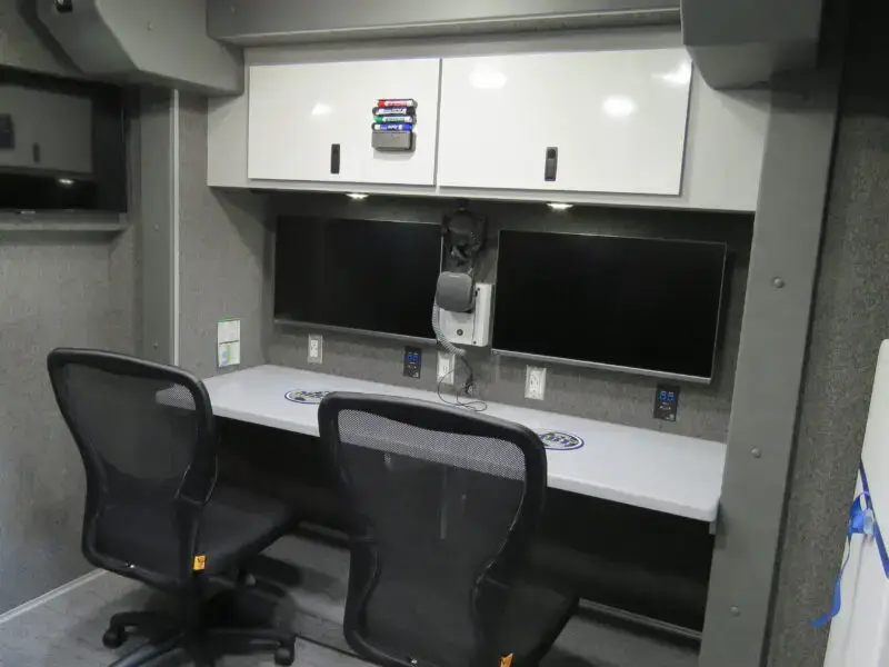 Two black office chairs are placed in front of a desk with two computer monitors, a keyboard, a microphone, and white overhead cabinets in a gray, modern workspace with a wall-mounted TV. Aurora Police Mobile Command