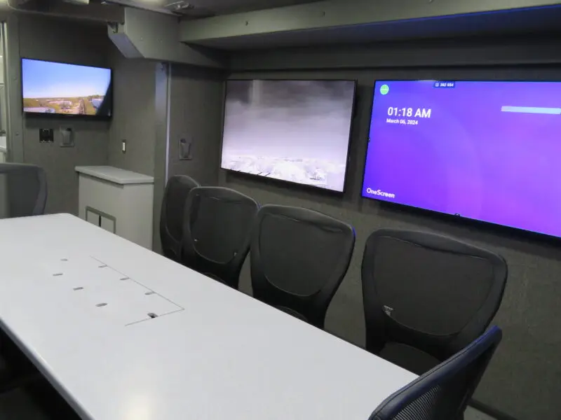 A modern conference room with a long white table, several black chairs, two large wall-mounted screens, and a smaller screen displaying security camera footage. The room has a gray color scheme and built-in cabinets.