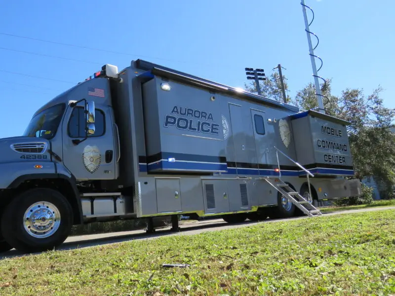 A large Aurora Police mobile command center vehicle is parked on a paved area near grass and trees. The vehicle has multiple compartments, stairs, and a raised antenna system. Aurora Police Mobile Command