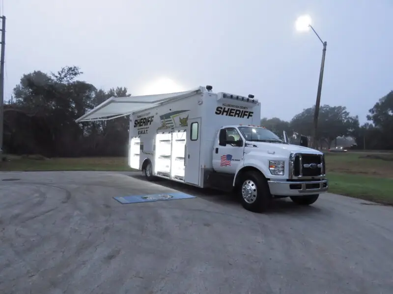 A large white sheriff’s mobile command vehicle is parked on a paved area at dusk with storage compartments open and an awning extended. Trees and grass are visible in the background.