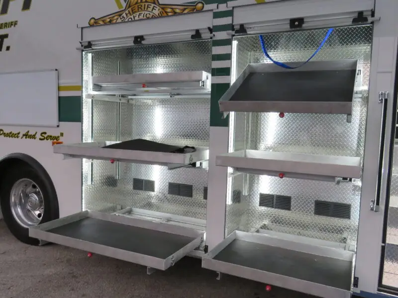 The image shows the side of a sheriff SWAT vehicle with multiple open metal compartments and pull-out trays, designed for organized storage of equipment. The trays and compartments are empty.