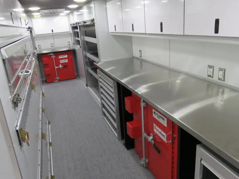 A clean, organized workspace with stainless steel countertops, white cabinets, drawers, and red tool chests along both walls, with bright overhead lighting. The floor is covered with gray carpet.