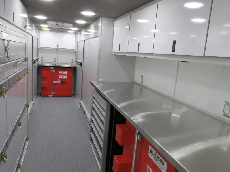A view inside a mobile workspace or service vehicle with metal cabinets, drawers, and work surfaces along the walls, overhead lighting, red storage units, and grey carpeted flooring.
