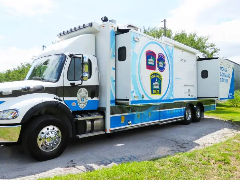 A large white and blue City of Coral Gables Mobile Command vehicle is parked on a road near grass and trees, displaying police logos and text indicating it is for emergency and community use. Coral Gables Mobile Command.