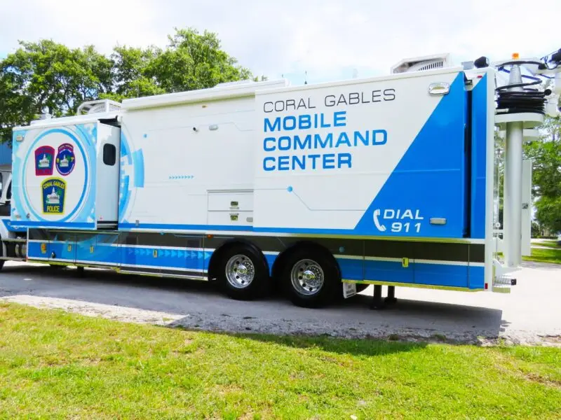 A large white and blue Mobile Command truck labeled Coral Gables is parked on a grassy area near trees and buildings; various law enforcement logos representing the city are displayed on its side. Coral Gables Mobile Command.