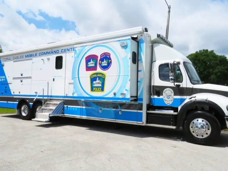 A large white and blue Mobile Command truck marked “Coral Gables Police” is parked on pavement near grass and trees under a partly cloudy sky. Coral Gables Mobile Command.