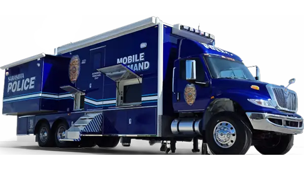 A Sarasota Police Mobile Command vehicle, painted blue with white stripes and official insignia, is parked on-site. It features an extended side compartment and stairway for entry, serving as a mobile command center for operations.