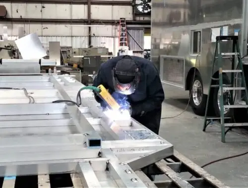 A person wearing protective gear is welding metal components in an industrial workshop, surrounded by large metal structures, tools, and equipment.