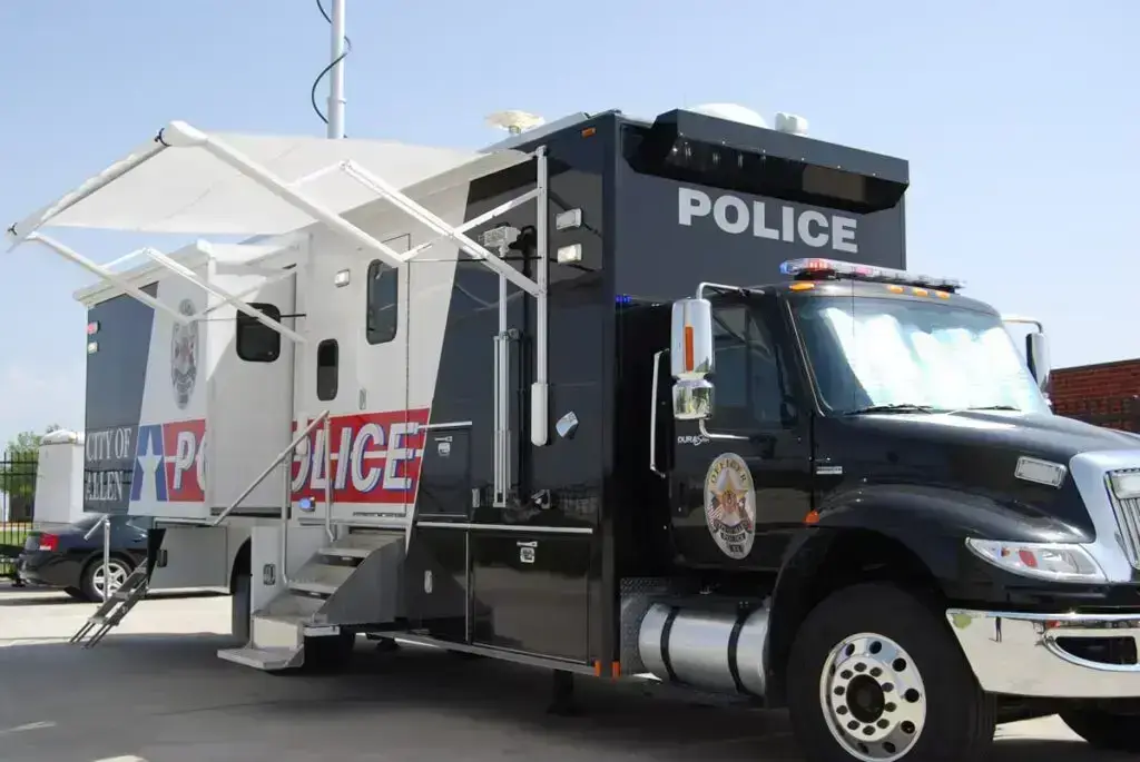 A large black and white police mobile command center truck, freshly finished with vehicle detailing, is parked on a concrete surface with its side awning extended. The vehicle features police insignia, markings, and exterior equipment.