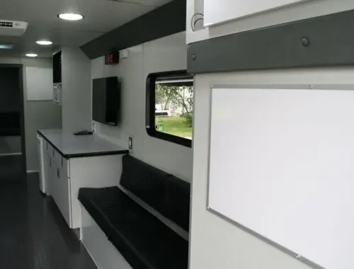 Interior of a mobile office or command center with a long bench, countertop, wall-mounted TV, storage cabinets, and a large blank whiteboard, featuring clean, modern surfaces and overhead lighting.