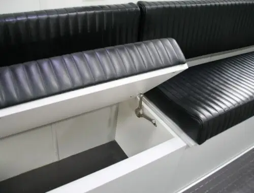 A close-up view of a black, vinyl-upholstered bench seat with the seat lifted to reveal a white storage compartment underneath. The lid is propped open by a metal support hinge.