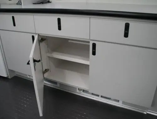 A white cabinet with black handles is shown with one lower door open, revealing two empty shelves inside. The cabinet is part of a set on a black tiled floor with a white countertop above.