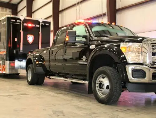 A black Ford pickup truck with emergency lights tows a large black trailer with markings inside a spacious warehouse with concrete floors and exposed beams. Rehab Unit Trailer