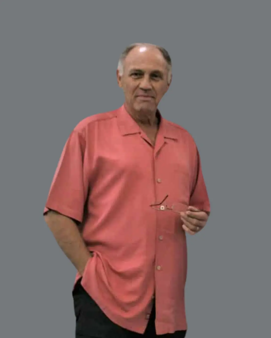 A man wearing a short-sleeved, coral-colored button-up shirt stands against a plain gray background, holding eyeglasses in his right hand and resting his left hand in his pocket.