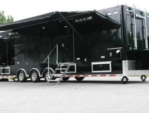 A large black mobile trailer with an extended awning, multiple windows, three axles, and metal stairs leading to an entrance, parked on a paved surface with grass and trees in the background. Rehab Unit Trailer