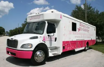 A large white and pink mobile clinic vehicle labeled Womens Center for Radiology offers 3D mammography and ultrasounds for screenings. The truck is parked on a paved surface with trees and a blue sky in the background. Women’s Center for Radiology