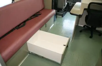 A white pull-out storage drawer is open beneath a maroon bench seat in a vehicle interior. The floor is green with a textured pattern. A table and office chair are seen nearby. Women’s Center for Radiology