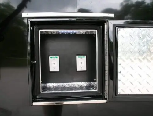 A close-up of an open metal compartment with a diamond plate door, revealing two labeled electrical switches mounted inside on a black panel. The compartment is part of a larger black structure outdoors. Rehab Unit Trailer