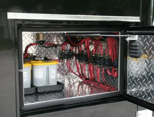 An open metal compartment displays batteries and multiple red cables connected to labeled switches and electrical components, all mounted on a diamond plate background. Rehab Unit Trailer