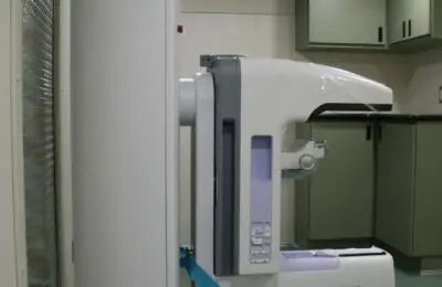 A medical imaging machine, specifically a mammography unit, is positioned in a clinical room with green flooring, green cabinets, and overhead lighting.