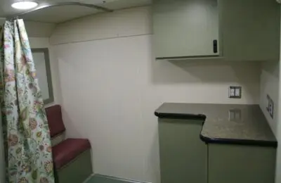 A small room with green cabinets, a gray countertop, two maroon cushioned seats, a floral curtain, and electrical outlets on a cream-colored wall. The decor appears simple and functional.