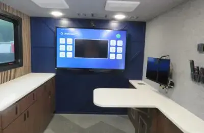 A modern mobile office interior with a large wall-mounted display screen showing a menu interface, a curved white desk, wood cabinetry, a window, a smaller monitor, and several remote controls mounted on the wall. University Federal Credit Union