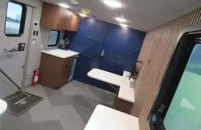 Interior of a modern RV or trailer with blue double doors, wood cabinets, white countertops, a coffee machine, a fire extinguisher, fold-out tables, carpeted floor, and a window showing greenery outside.