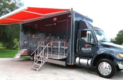 A large black and gray mobile office truck with an extended bright orange awning, metal steps leading to the entrance, and company logos on the side, is parked on a paved area near grass and trees. Granite Credit Union