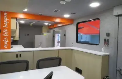 Interior of a modern mobile office or medical unit with gray walls, white countertops, light wood cabinets, mesh chairs, and an orange sign reading “make life happen” along an upper beam.