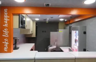 Interior of a modern mobile office or RV with white counters, light wood cabinets, a microwave, a coffee maker, and an orange beam displaying the text make life happen in white letters.