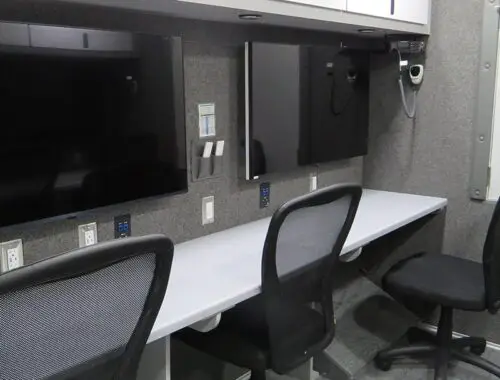 A workspace with three office chairs at a long desk, two large wall-mounted monitors, white overhead cabinets, power outlets, and a gray carpeted floor. The setting appears to be inside a mobile unit or trailer. Bellevue Trailer Command
