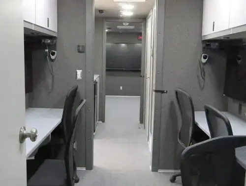 The image shows the interior of a mobile office or command center with desks, office chairs, overhead cabinets, and computer monitors lined along gray carpeted walls. The area is well-lit with recessed ceiling lights.
