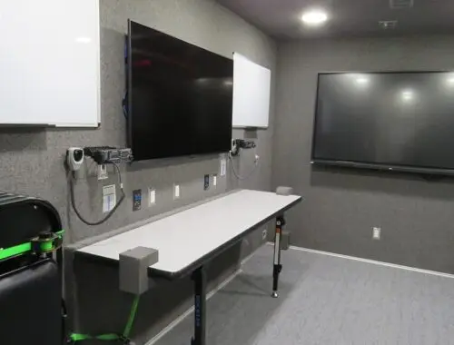 A small room with gray walls containing two folding tables, one large wall-mounted TV screen, a smaller TV screen, whiteboards, a digital clock, and stacked folding chairs in the corner.