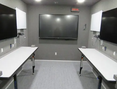 A room with two long tables along opposite walls, large flat-screen monitors above each table, a central wall-mounted monitor, whiteboards, electrical outlets, and a digital clock displaying the time 09:46.