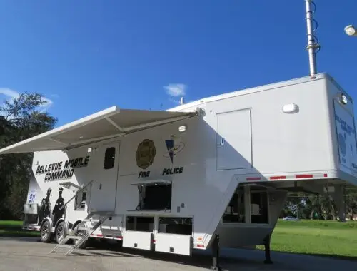 A large white mobile command center trailer labeled Belleview Mobile Command, with police and fire logos, parked outdoors on pavement under a blue sky with some clouds, trees, and grass in the background. Bellevue Trailer Command