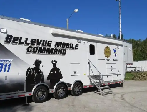 A large white trailer labeled Bellevue Mobile Command with graphics of emergency personnel, 911 signage, and logos for fire and police, parked on a paved area beside grass and trees under a clear blue sky. Bellevue Trailer Command