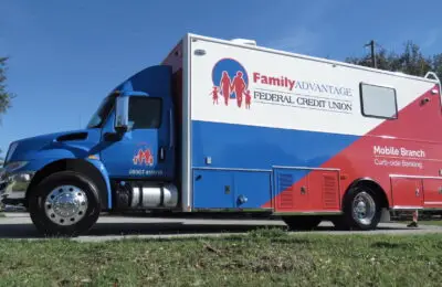 A Family Advantage Federal Credit Union mobile banking truck labeled “Mobile Branch Curb-side Banking” is parked outdoors on a sunny day, surrounded by trees and grass. Family Advantage Federal Credit Union