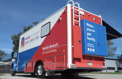 A large red, white, and blue mobile banking truck from Family Advantage Federal Credit Union features a rear door with the words Relax, youve got family. It is parked outdoors near a building and trees under a clear blue sky. Family Advantage Federal Credit Union