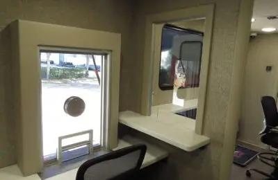 Interior view of a Family Advantage Federal Credit Union office, featuring a glass window with a circular opening, a counter ledge, chairs, and a door in the background. A window reveals a parked vehicle and trees outside.