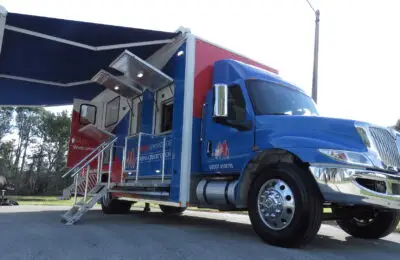 A large blue and red Credit Union mobile unit with extended awning and stairs is parked on a paved area. The side door is open, revealing an entrance ramp and metal railings. Trees and a chair are visible in the background. Family Advantage Federal Credit Union
