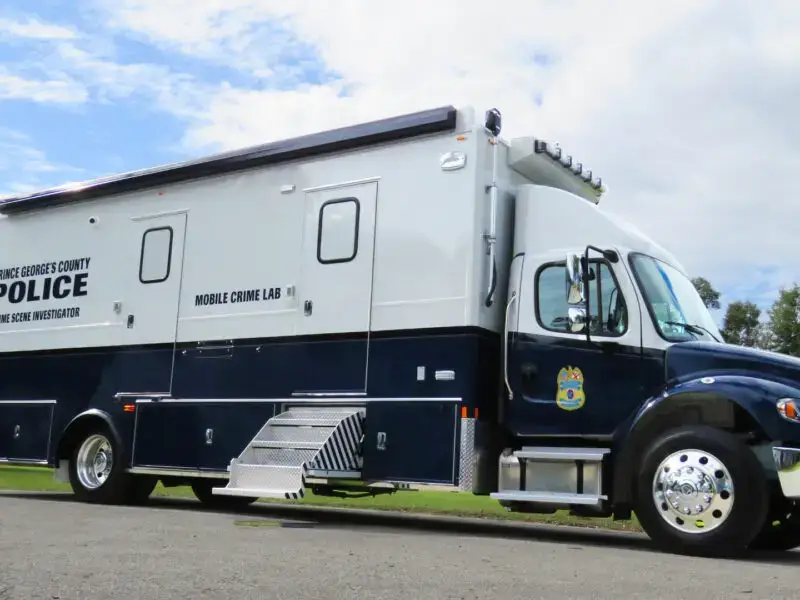 A large Prince Georges County Crime Scene Vehicle is parked outdoors on a paved surface, with trees and a partly cloudy sky in the background.