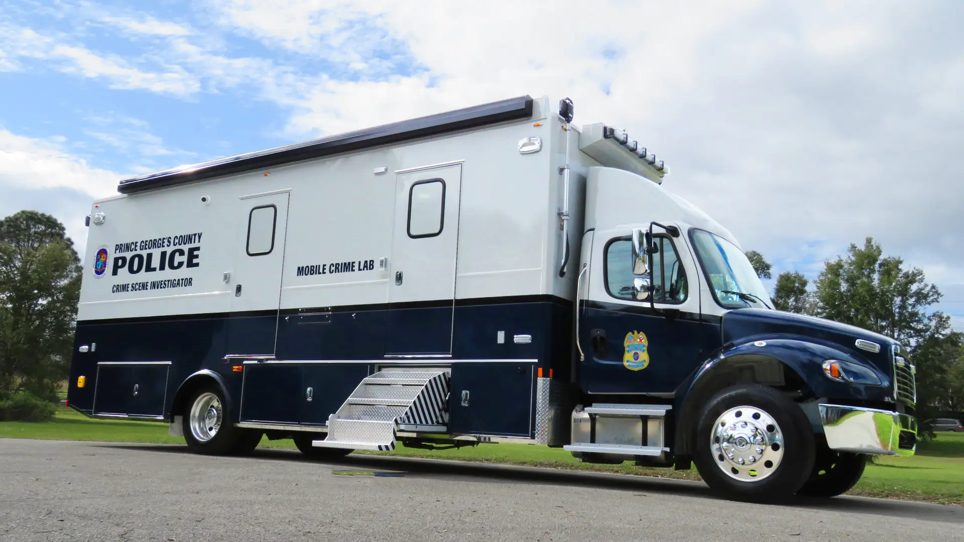 A large mobile crime lab vehicle belonging to the Prince George’s County Police is parked outdoors on pavement, featuring blue and white colors, police branding, and multiple compartments.