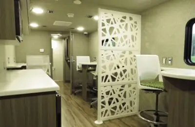 Interior of a modern RV or mobile office designed for banking or financial services, featuring wood flooring, built-in counters, a decorative white room divider, mesh chairs at small tables, and a window on the right letting in natural light.