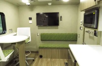 Small, modern RV interior with a wall-mounted TV, green-patterned bench, round table with two white chairs, microwave, cabinets, and window on the left. The wood-patterned vinyl floor creates a cozy space—perfect for discussing Credit Union 1 financial services.