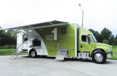 A large green and white Credit Union 1 mobile truck is parked on pavement, with stairs leading to its open side doors and awnings extended, ready to offer financial services. Trees and grass are visible in the background.