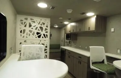A modern RV interior features a kitchenette with wood cabinets, a white countertop, and built-in appliances, alongside a dining area with white chairs and a decorative white partition—perfect for Credit Union 1 members on the go.