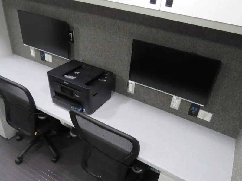 Two office chairs are placed at a long desk with two computer monitors, a black printer in the middle, power outlets, and overhead cabinets. The workspace, inspired by a Crime Scene Vehicle in Prince Georges County, has a gray color scheme and minimal decor.